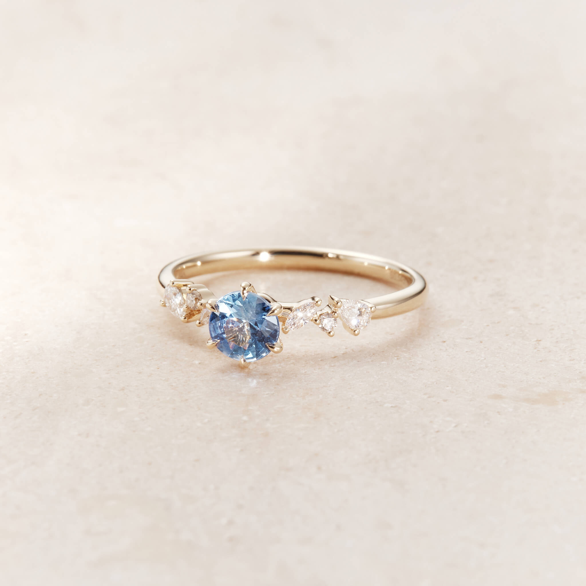 A close-up studio photograph showcases a delicate Yellow Gold ring resting angled forward on a textured, pale beige stone surface.