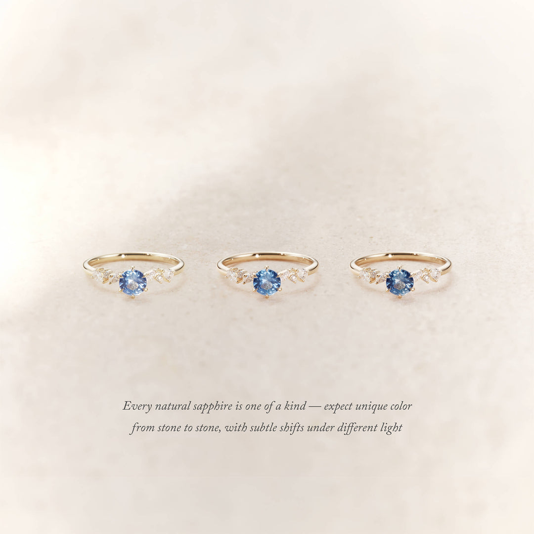 Three Yellow Gold rings with round light blue sapphires and small diamond accents on each side are displayed side by side on a soft beige background.