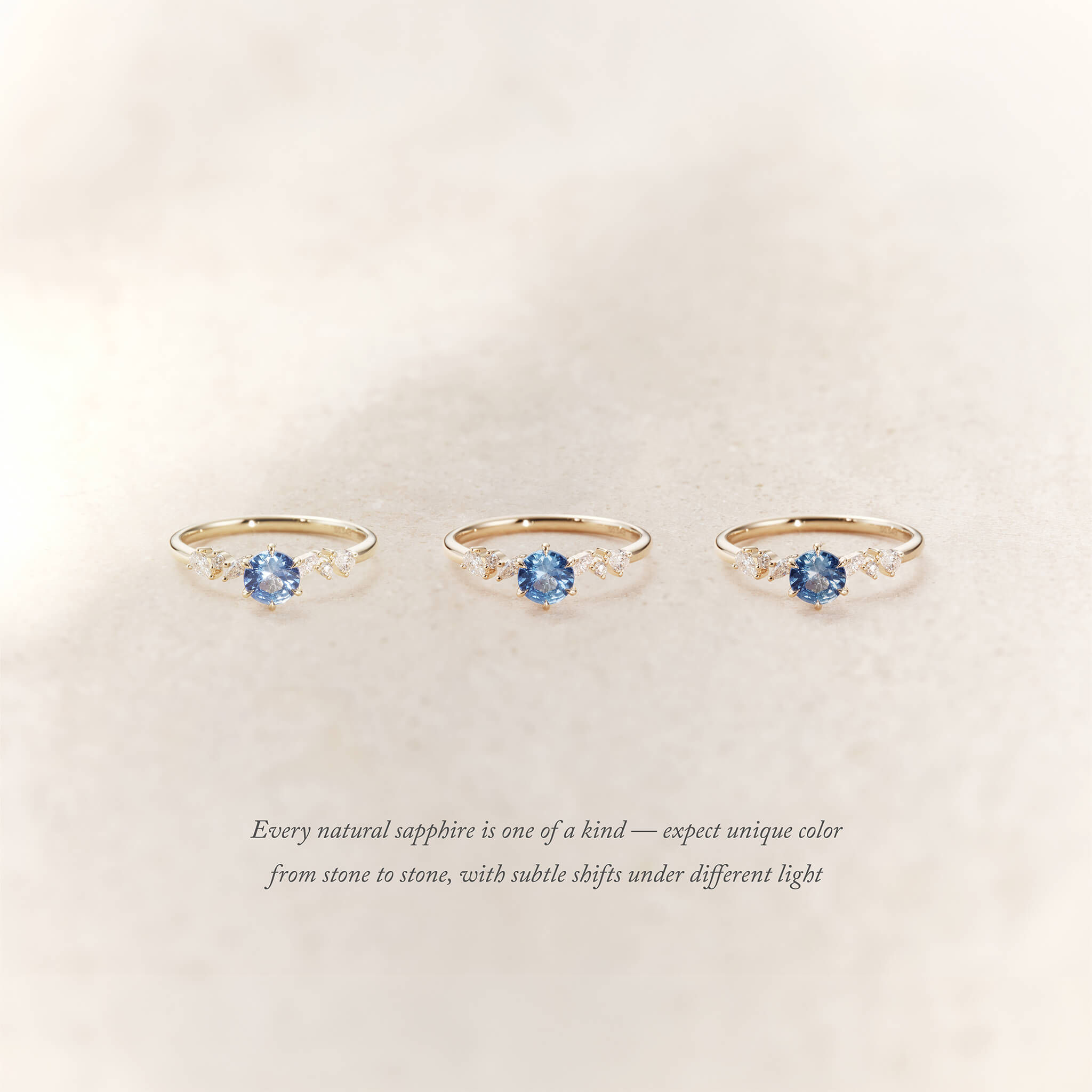 Three Yellow Gold rings with round light blue sapphires and small diamond accents on each side are displayed side by side on a soft beige background.