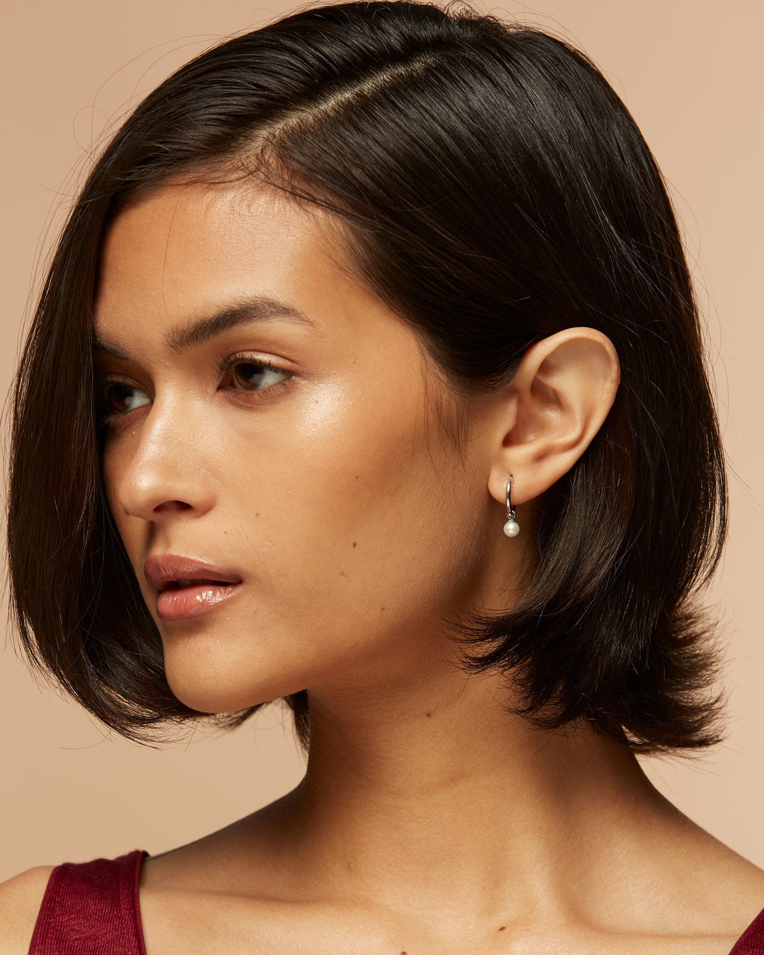 A person with short, dark hair and pearl earrings gazes sideways against a soft beige background, conveying a serene and thoughtful expression.