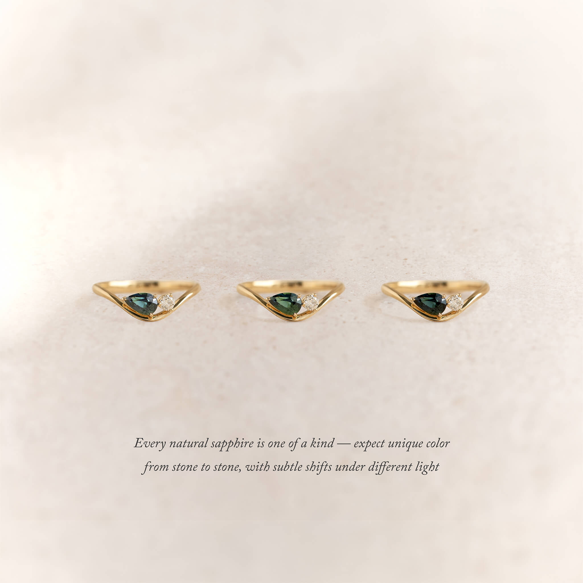 Three Yellow Gold rings with pear-shaped deep green sapphires and a small round diamond accent are displayed side by side on a soft beige background.