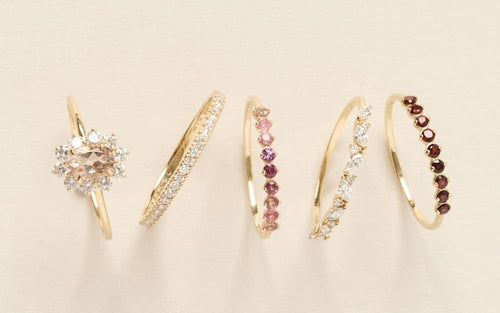 Close-up of five delicate gold rings featuring clear, pink, and red small round gemstones on a beige background