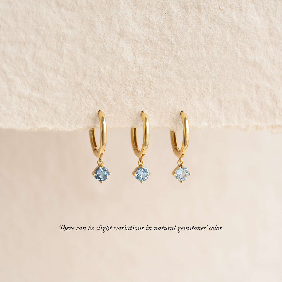 Three gold hoop earrings with small dangling light blue gemstones are displayed hanging from a soft, textured fabric background, with a note below indicating that natural gemstone colors may vary.