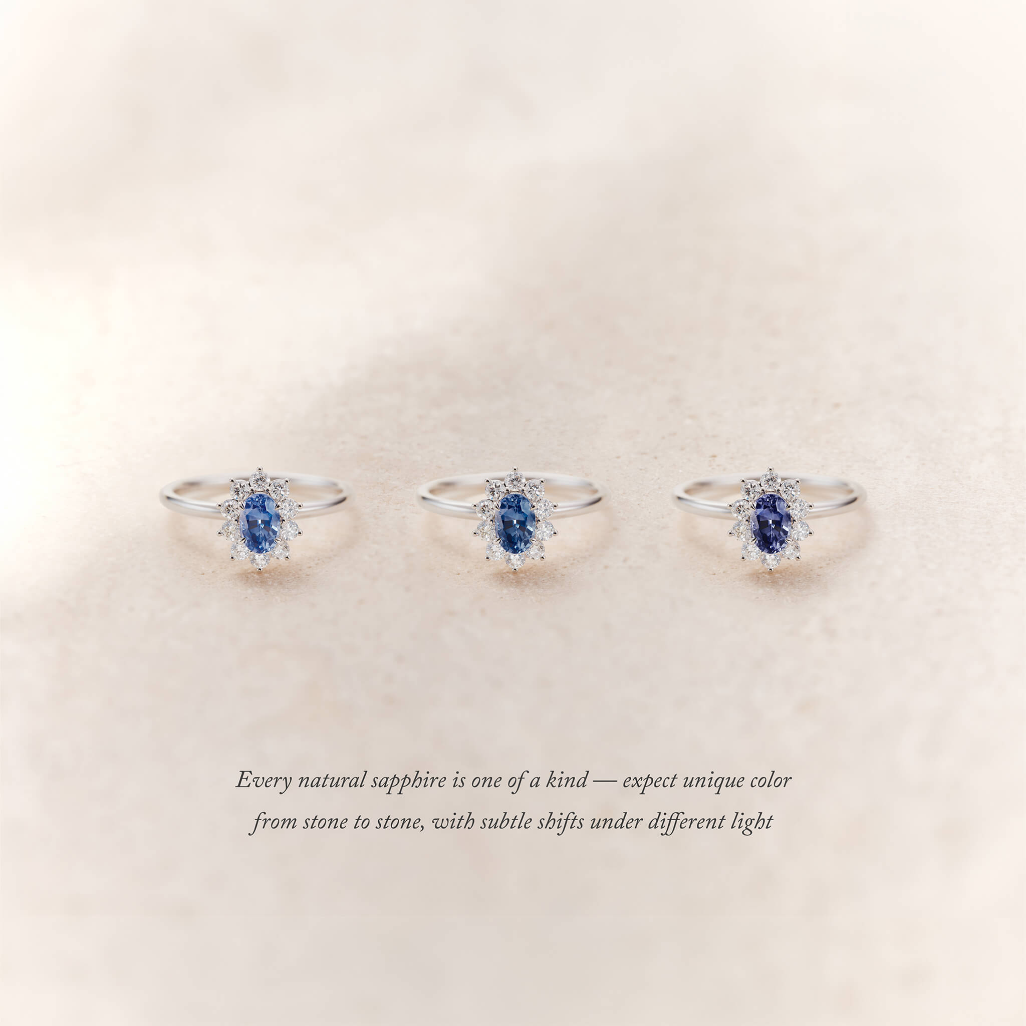 Three White Gold halo rings with oval blue sapphires surrounded by small diamonds are displayed side by side on a soft beige background.