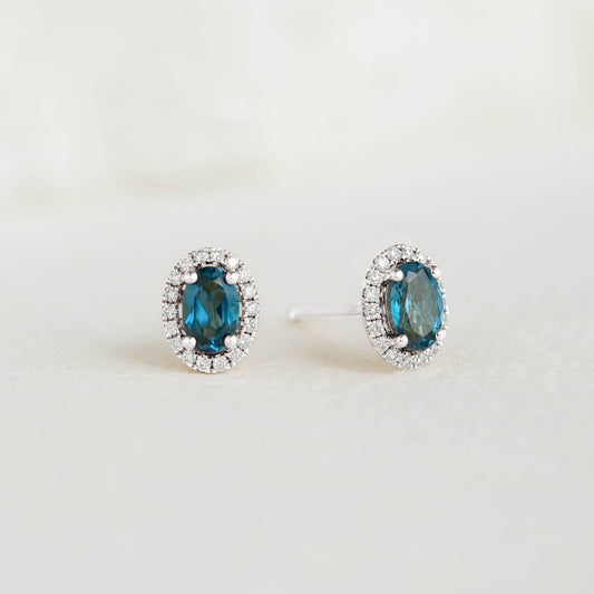 London Blue Topaz Halo Stud Earrings White Gold - Marzia with oval-cut blue topaz and diamond halo on a neutral background