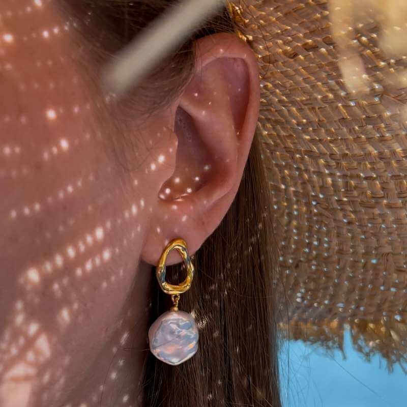 Close-up of an ear wearing a gold hoop earring with a dangling irregular pearl against a straw hat and blue sky background