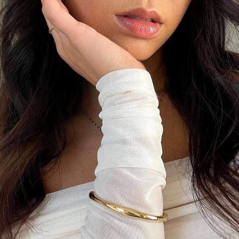 Close-up of a woman wearing a white long-sleeve top, gold bangle, and delicate gold ring with dark wavy hair visible