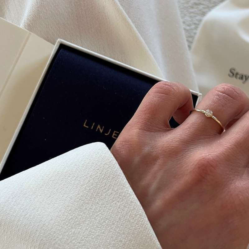 Close-up of a hand wearing a delicate gold ring with a small round clear stone over a white fabric and dark blue box background