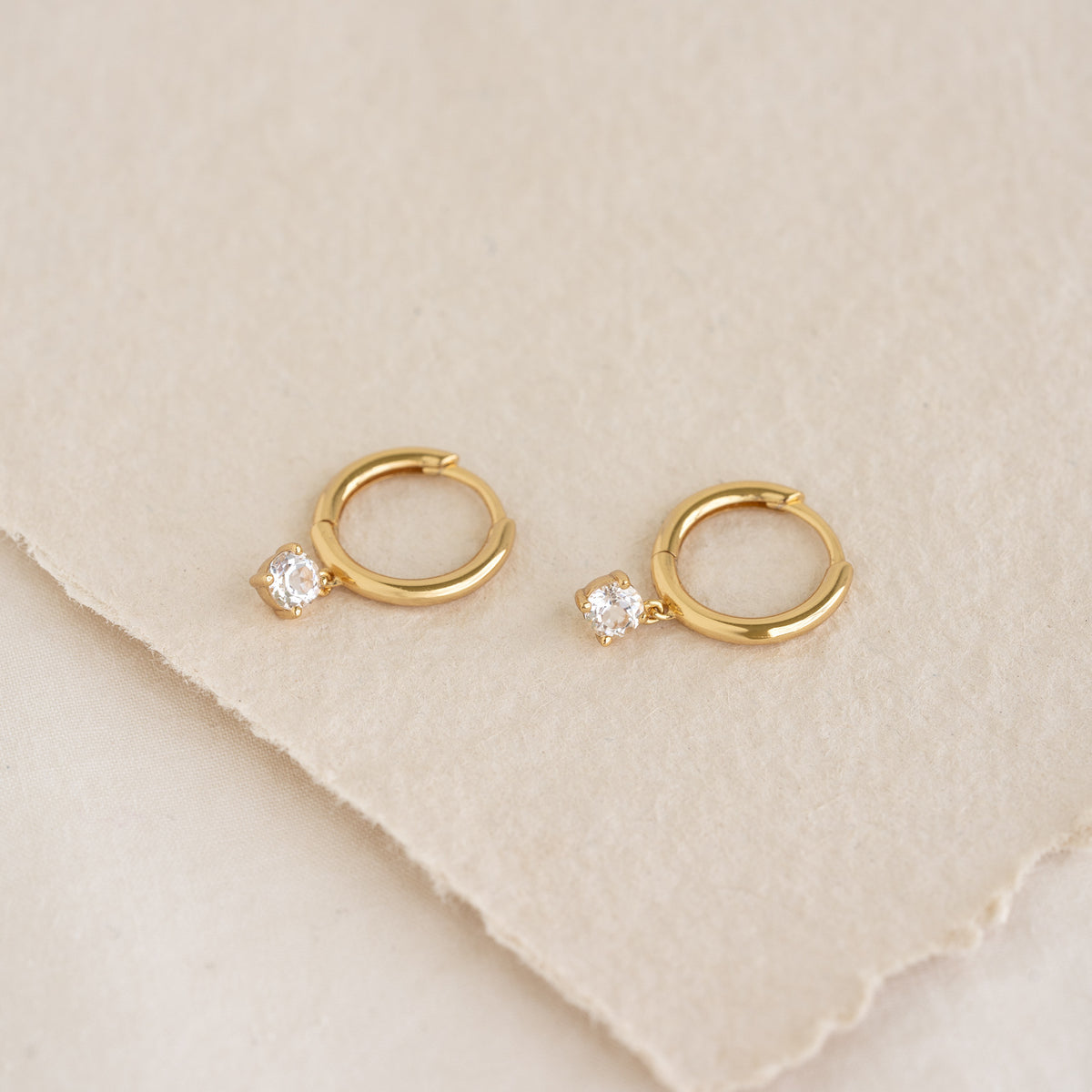 Close-up of April Birthstone Drop Huggie Earrings - White Topaz in gold vermeil on a neutral textured background