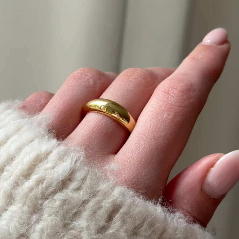 Close-up of a hand wearing a smooth polished gold band ring with a soft cream-colored knit sleeve background