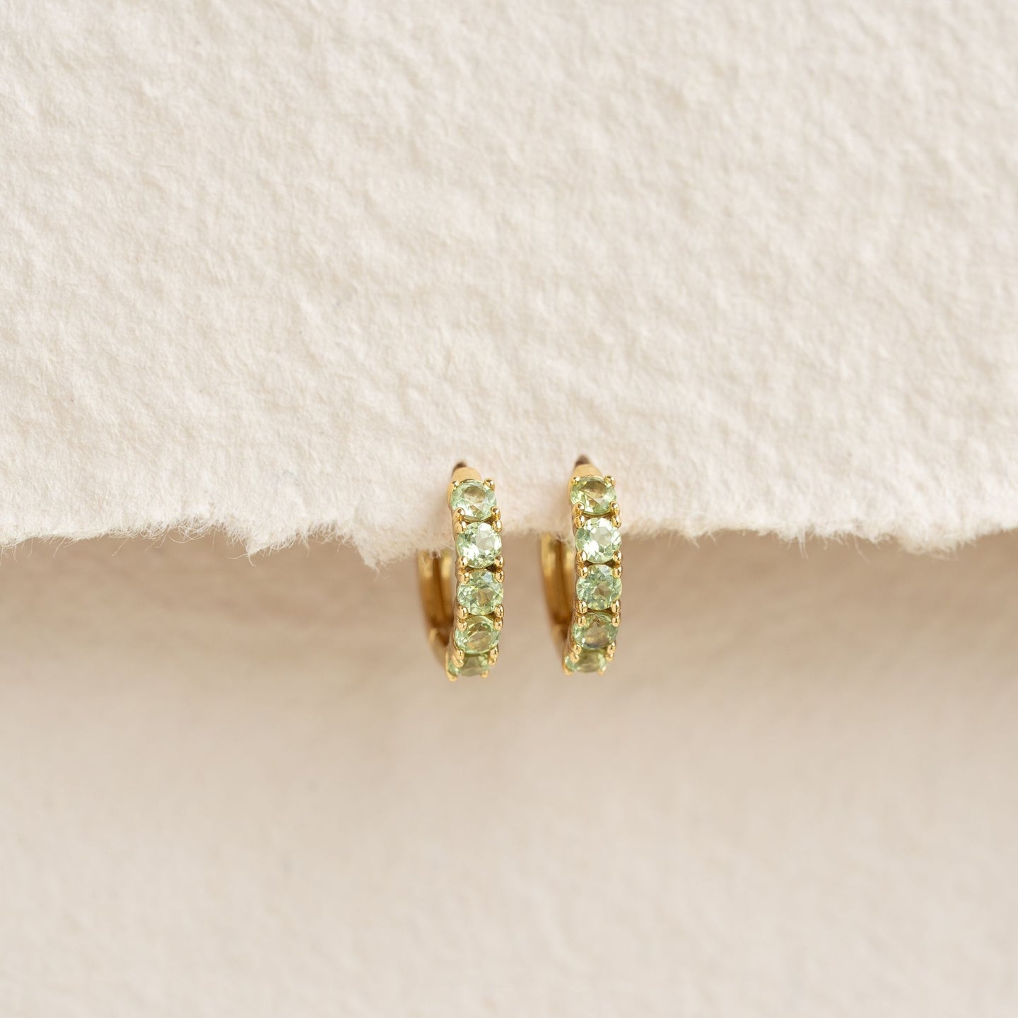 Close-up of August Birthstone Huggie Earrings - Olivine with green gemstones set in gold vermeil on a beige background