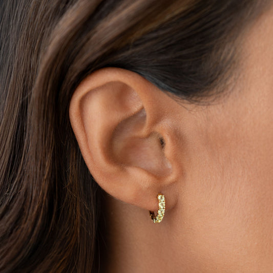 Close-up of an ear wearing August Birthstone Huggie Earrings - Olivine with gold vermeil setting on a neutral background
