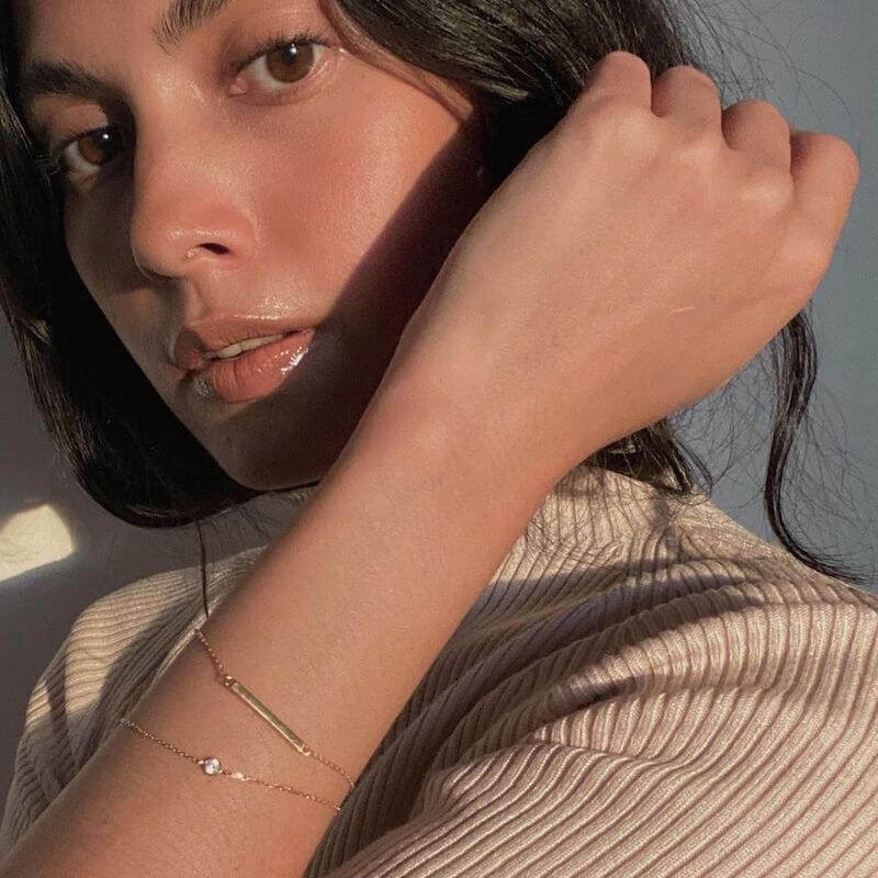 Close-up of a woman with natural makeup wearing a ribbed beige top and delicate gold bracelets in soft sunlight