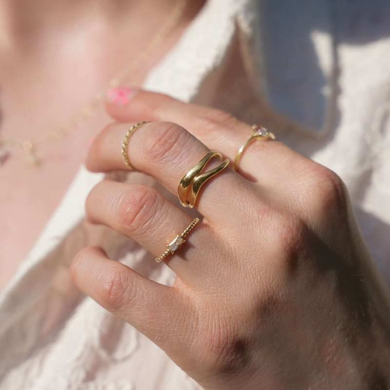 Close-up of a hand wearing three gold rings with different designs and a delicate gold necklace on a neutral background