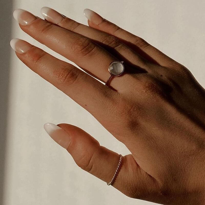 Close-up of a hand with long natural nails wearing a delicate silver ring with a round translucent stone on a neutral background