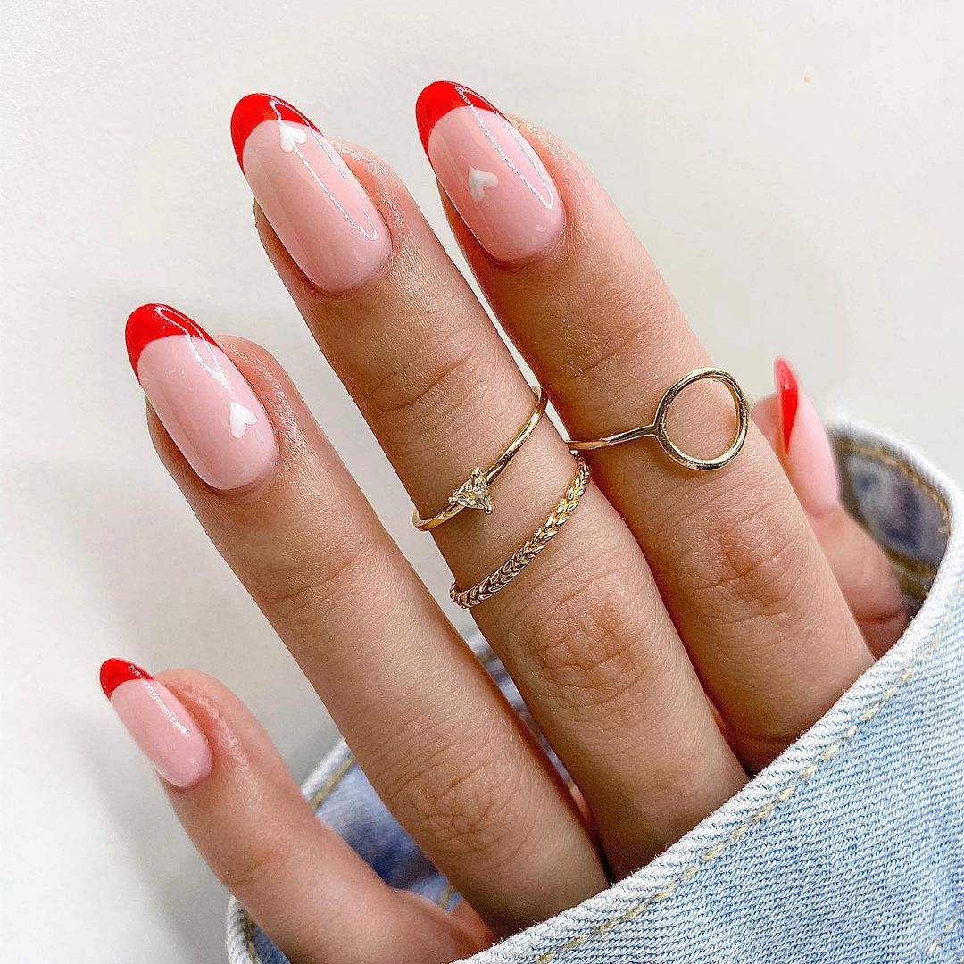 Close-up of a hand with nude nails and red French tips wearing gold rings and a denim jacket sleeve on a white background