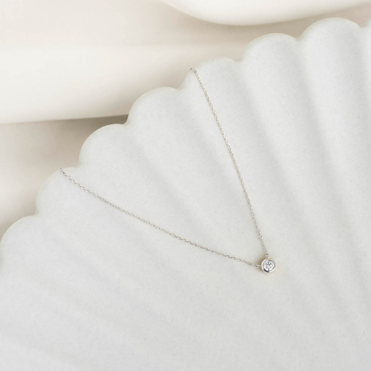 Bezel Set Diamond Necklace White Gold - Thais with a delicate chain and single bezel-set diamond on a white textured surface