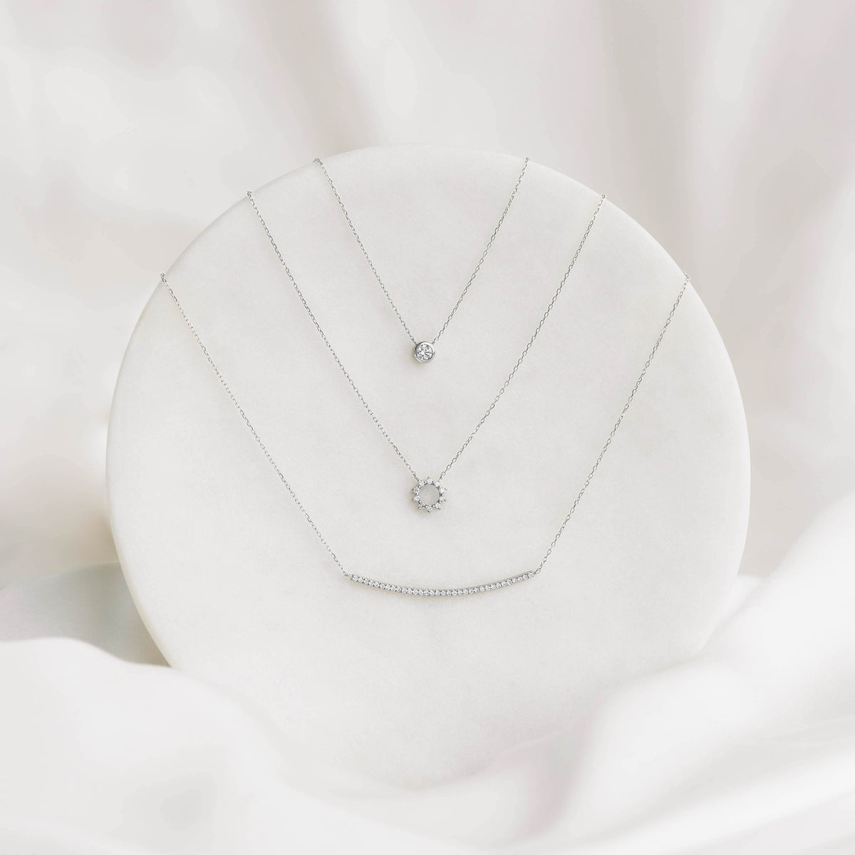 Three delicate white gold necklaces with bezel set diamonds on a round white display for Bezel Set Diamond Necklace White Gold - Thais