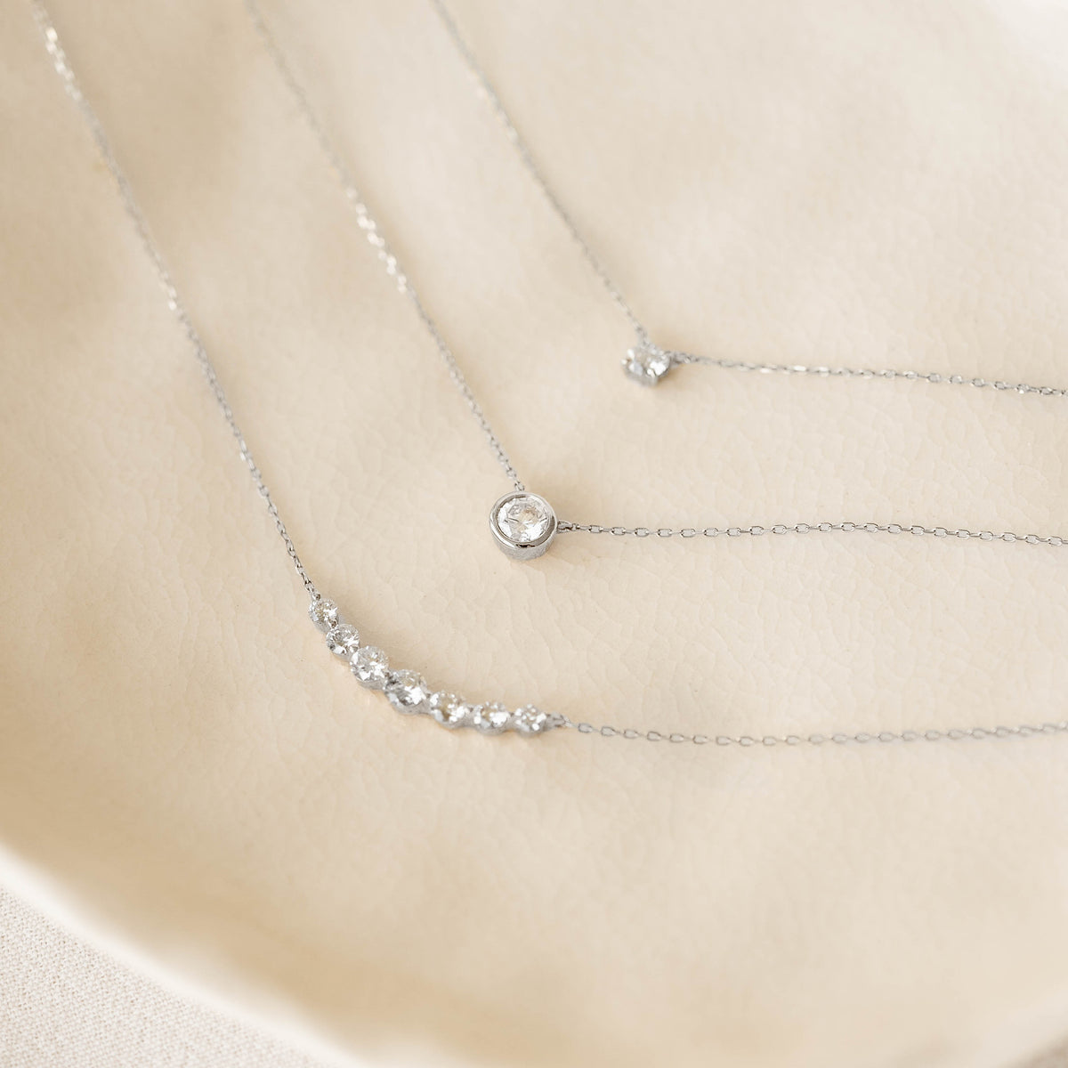 Close-up of Bezel Set Diamond Necklace 4mm White Gold - Ulla layered with delicate white gold chains on a soft beige background