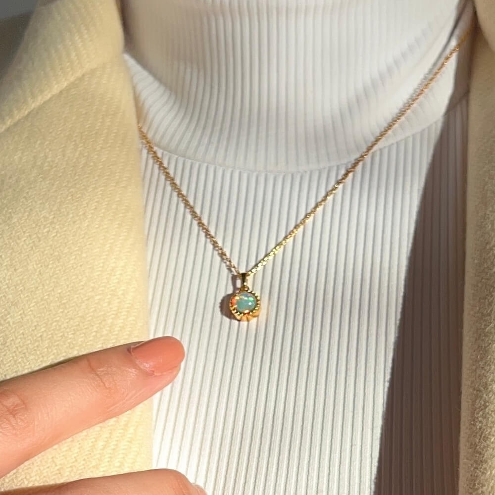 Close-up of a gold necklace with a round opal pendant worn over a white ribbed turtleneck and beige jacket