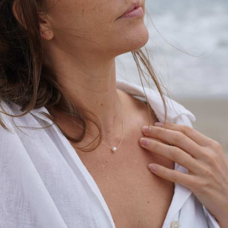 Close-up of a woman wearing a delicate gold necklace with a single pearl pendant against a blurred beach background