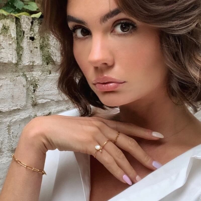 Close-up of a woman with natural makeup wearing gold rings and a bracelet with a white top against a textured brick wall