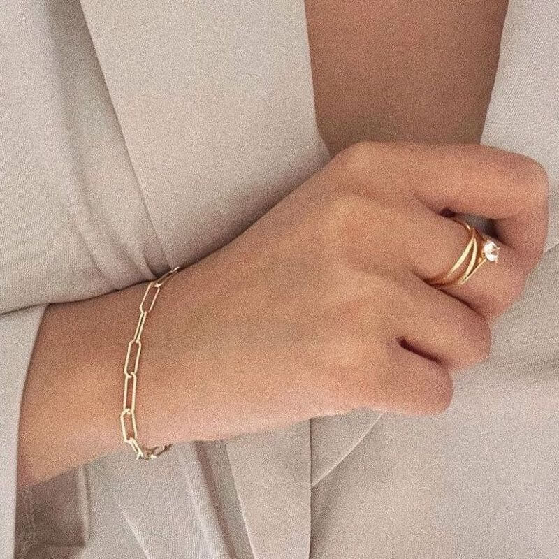 Close-up of a hand wearing a gold chain bracelet and two gold rings with a diamond on a beige fabric background