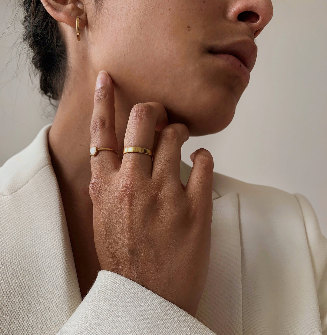 Close-up of a woman wearing small gold hoop earrings and two gold rings touching her neck in a white textured blazer