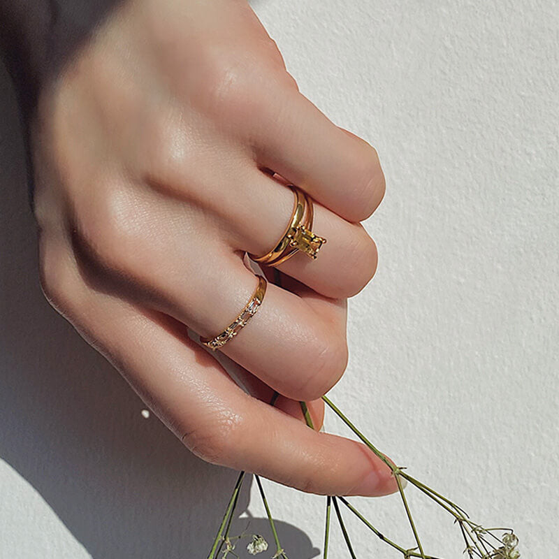 Close-up of a hand with two gold rings, one featuring a solitaire stone and the other with small embedded stones, holding delicate greenery