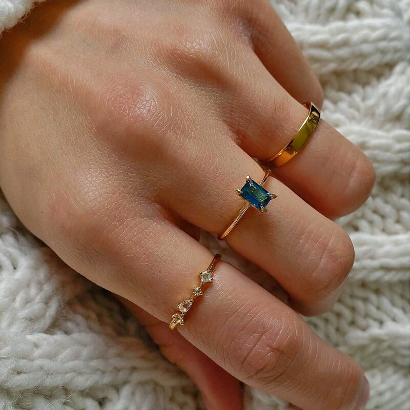 Close-up of a hand resting on a knitted fabric wearing three delicate gold rings, including one with a rectangular blue stone