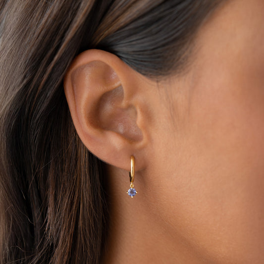 Close-up of ear wearing December Birthstone Drop Huggie Earrings - Cordierite with gold vermeil setting on neutral background