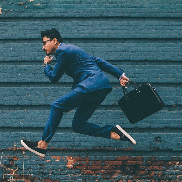 Man in a blue suit and black sneakers jumping mid-air against a blue textured wall carrying a black briefcase