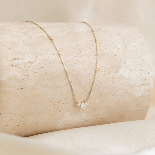 Elegant Gold necklace with three pear-shaped diamonds on a smooth beige stone surface, creating a luxurious and minimalist aesthetic.