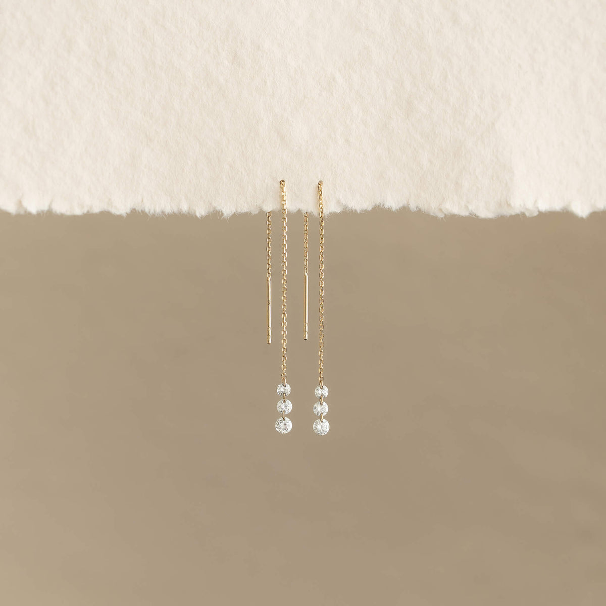 Gold threader earrings with delicate chains and sparkling crystals hang elegantly against a soft beige background, conveying simplicity and elegance.
