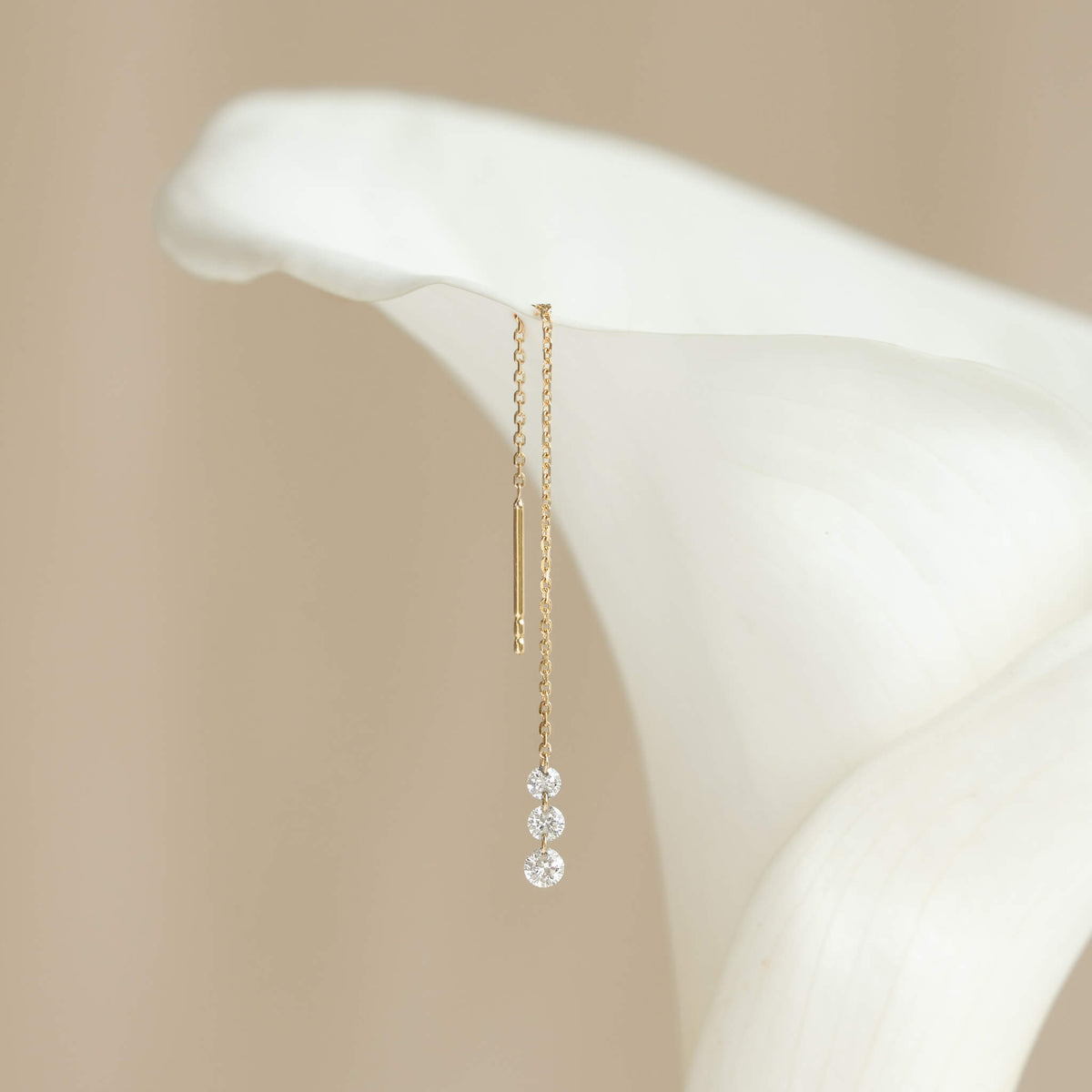 Elegant Gold earring with three diamonds hangs on a white calla lily against a soft beige background, conveying a sense of delicate luxury and serenity.