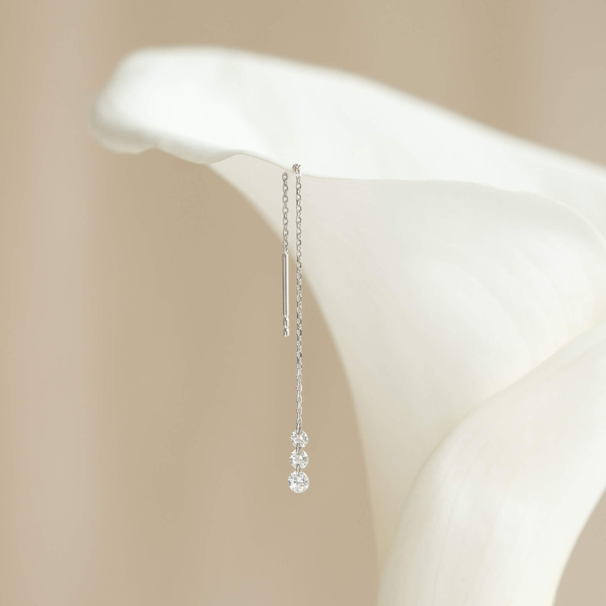 Elegant White Gold earring with three dangling diamonds hanging from a white calla lily petal, set against a soft beige background, evoking a delicate and serene mood.