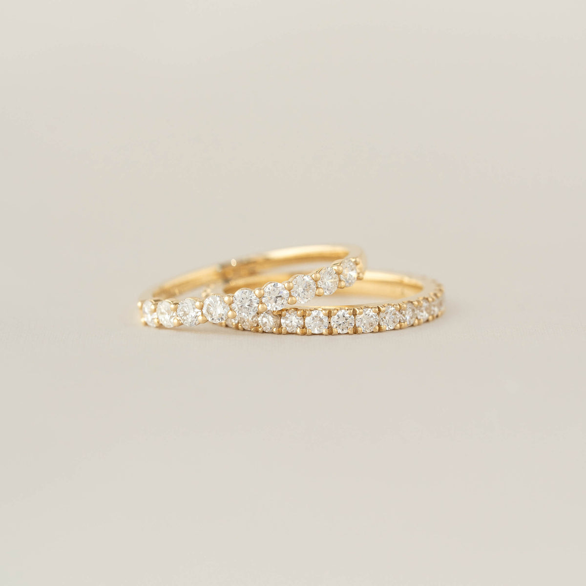 Two Gold rings adorned with small, sparkling diamonds are stacked against a soft beige background, conveying elegance and simplicity.