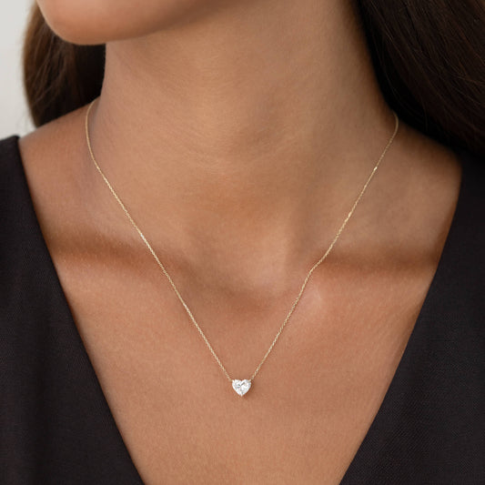 A close-up portrait of a woman with dark hair wearing a delicate Gold chain necklace featuring a small, brilliant-cut diamond shaped like a heart, paired with a simple dark V-neck top.