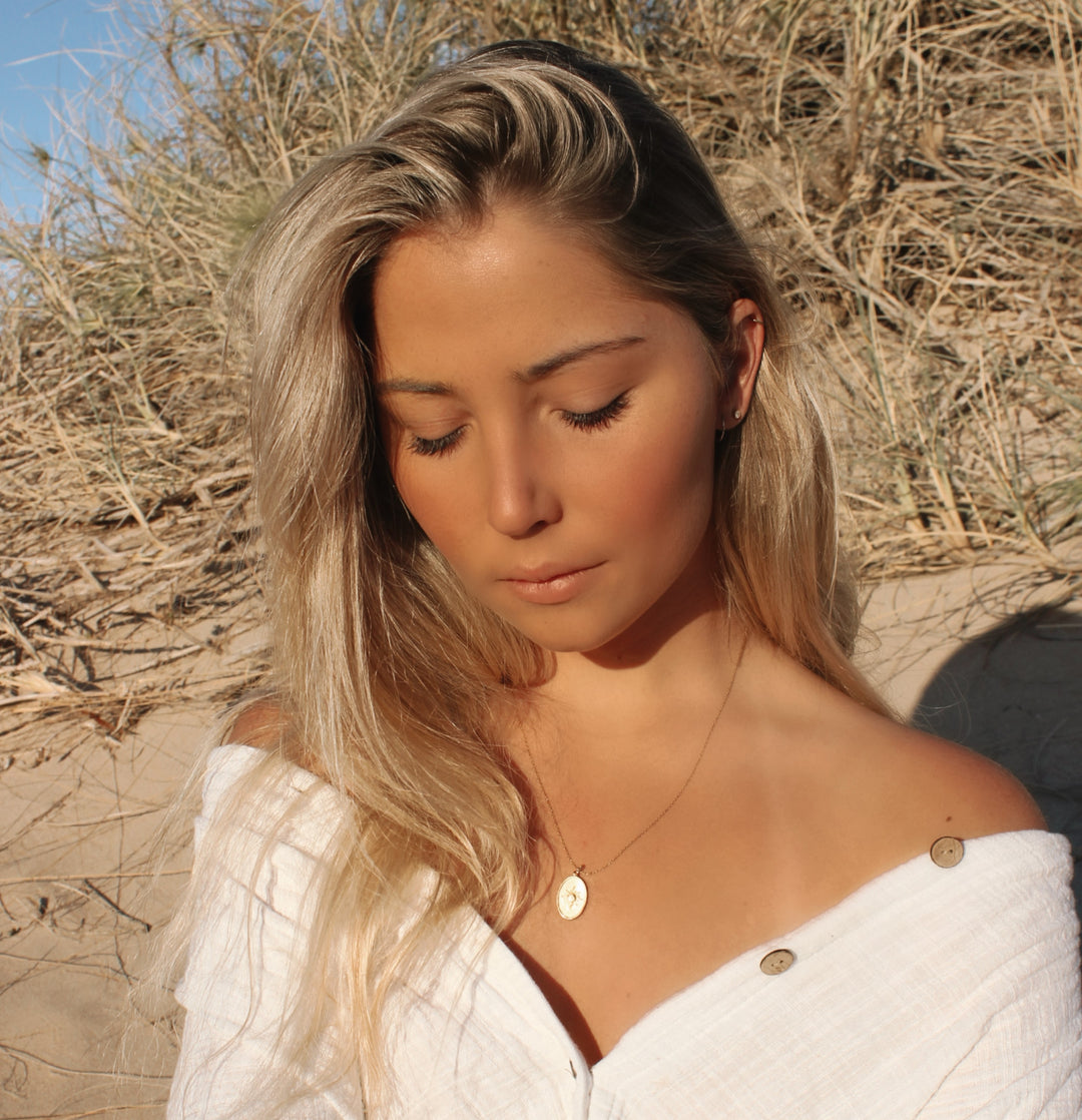 Young woman with long blonde hair wearing a white off-shoulder top and gold pendant necklace outdoors in natural light