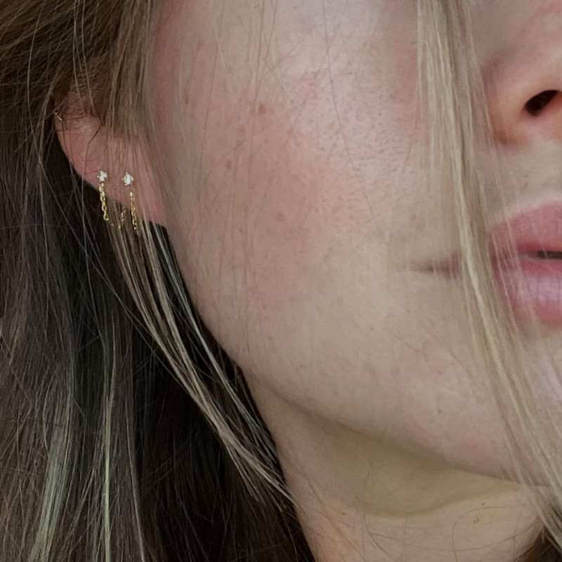 Close-up of a woman’s ear with three small gold earrings featuring delicate chain details against light skin and hair strands