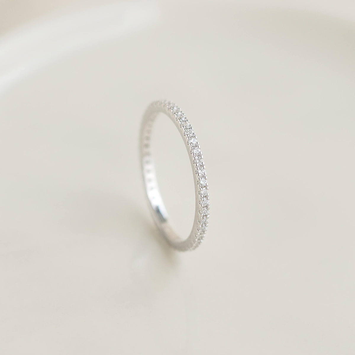 A delicate White Gold diamond eternity band with small sparkling stones set in a thin circle on a soft, neutral background, exuding elegance and simplicity.