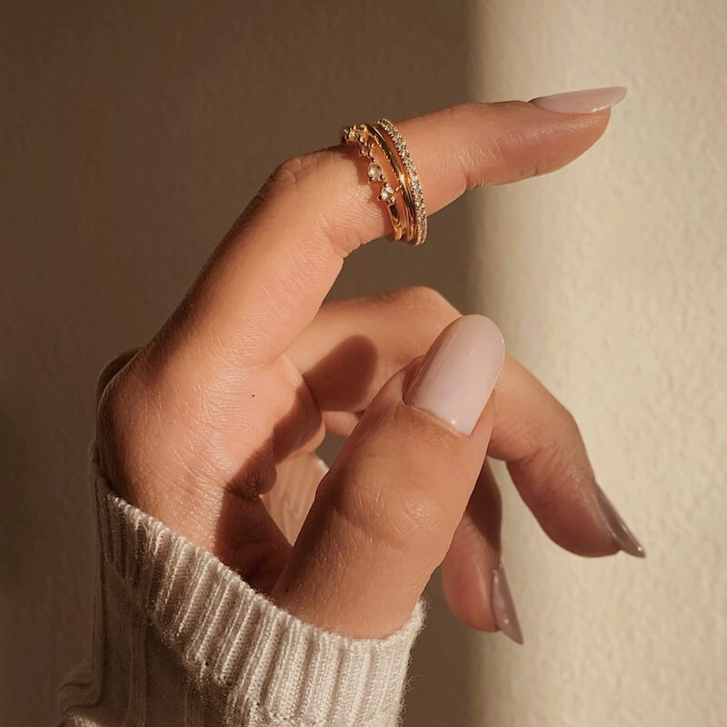 Close-up of a hand with light beige nail polish wearing stacked gold rings against a beige background