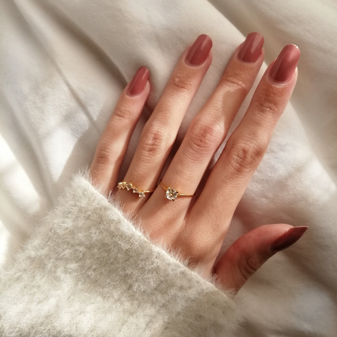 Close-up of a hand with mauve nail polish wearing two delicate gold rings resting on a soft white fabric background