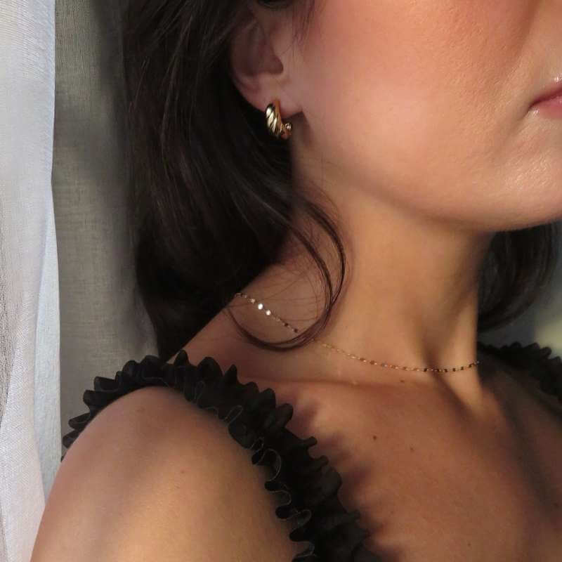 Close-up of a woman wearing a small twisted gold hoop earring and a delicate gold chain necklace with a ruffled black strap visible