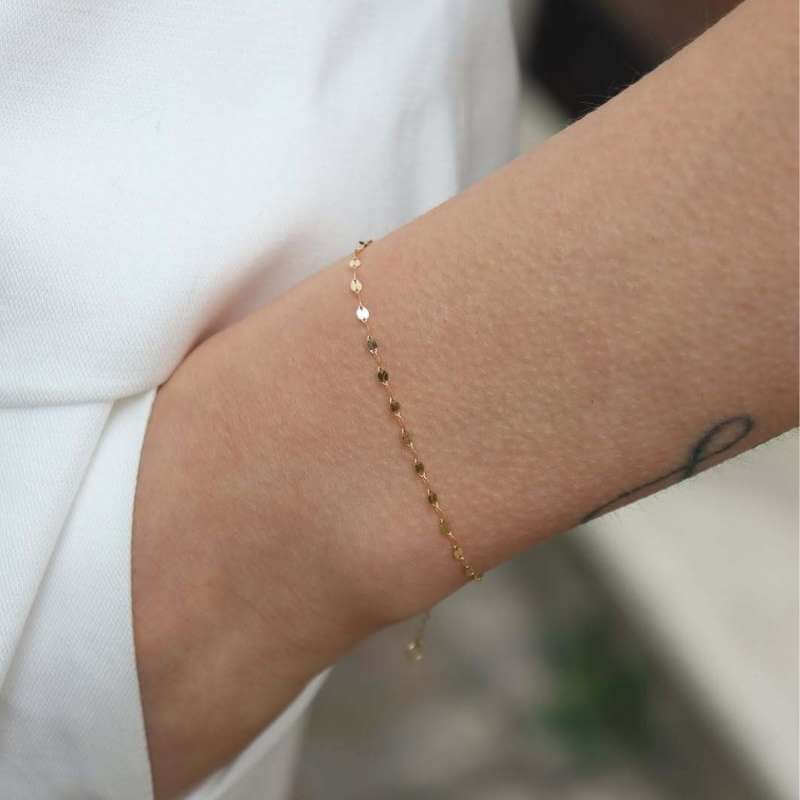 Close-up of a delicate gold chain bracelet on a wrist with a small tattoo, set against white fabric background