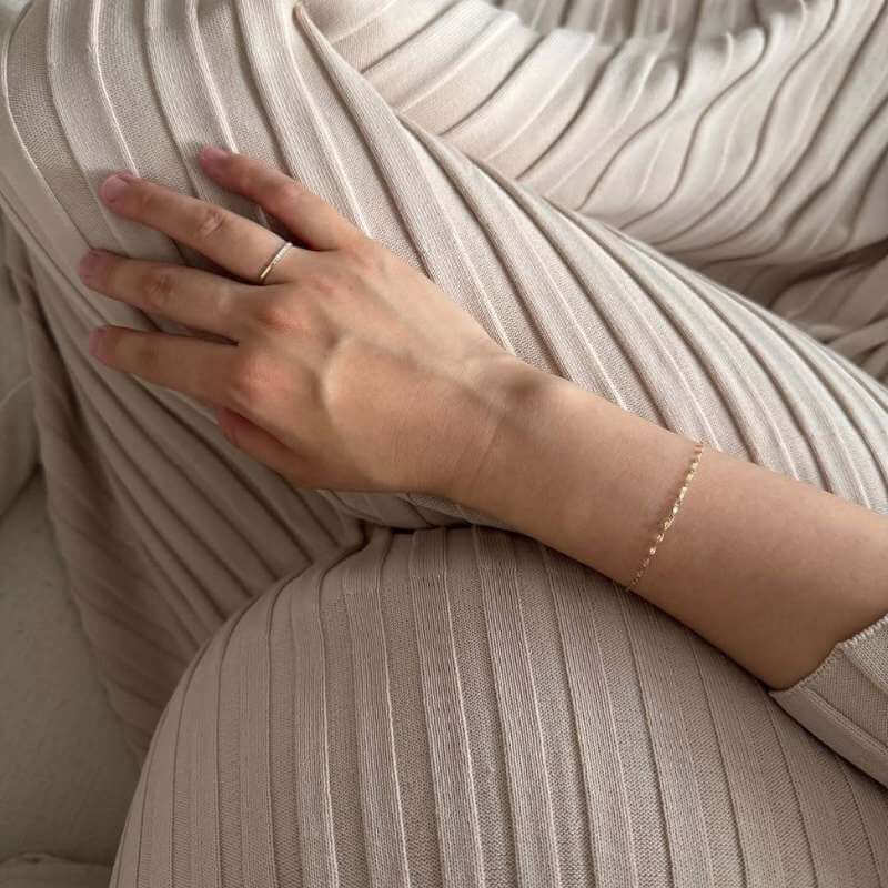 Close-up of a hand resting on beige ribbed fabric wearing a delicate thin gold bracelet and simple gold ring