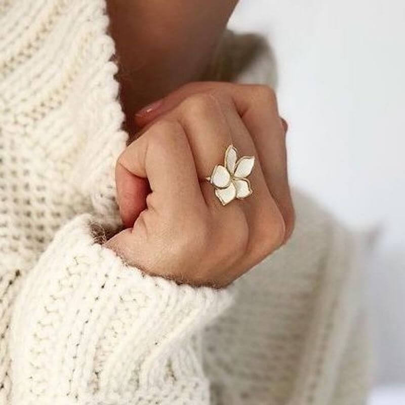Close-up of a hand wearing a white floral ring and a cream knitted sweater against a neutral background