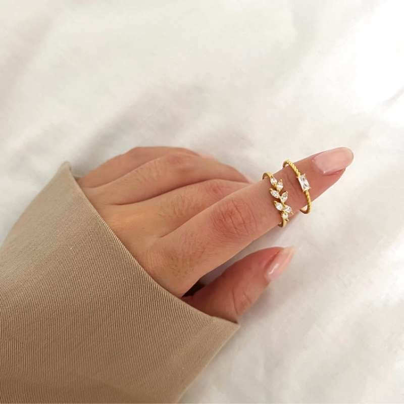 Close-up of a hand with manicured nails wearing two delicate gold rings with clear stones on a white fabric background