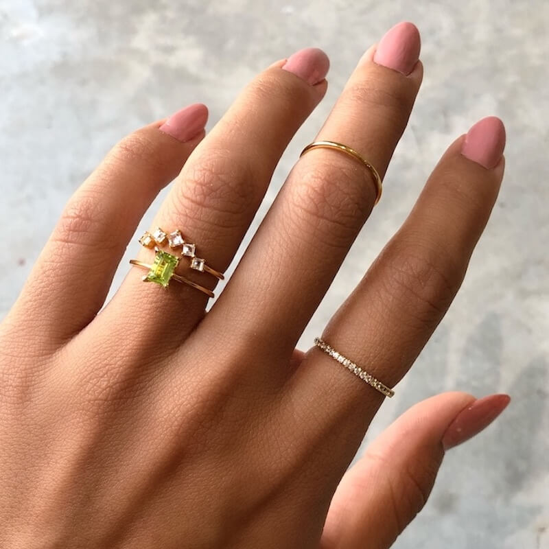 Close-up of a hand with pink nails wearing three delicate gold rings and one silver ring on a neutral background