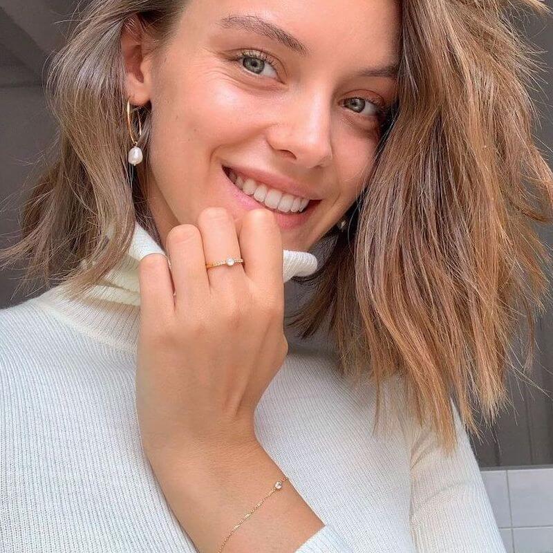 Close-up of a smiling woman with light brown hair wearing a white turtleneck, delicate gold bracelet, and pearl earrings